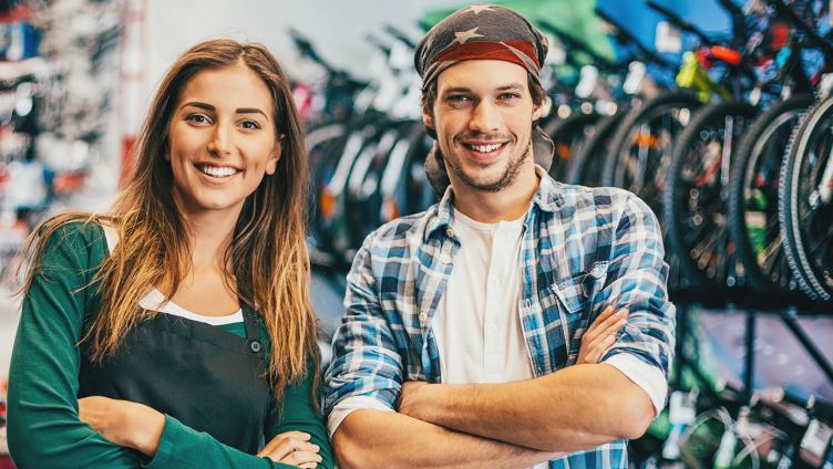 A young woman and a young man stand in the bicycle shop and smile in the camera. 