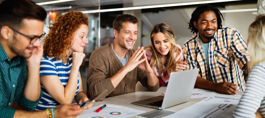 Lächelnde junge Menschen stehen um einen Tisch, auf dem ein Laptop steht und Unterlagen liegen.