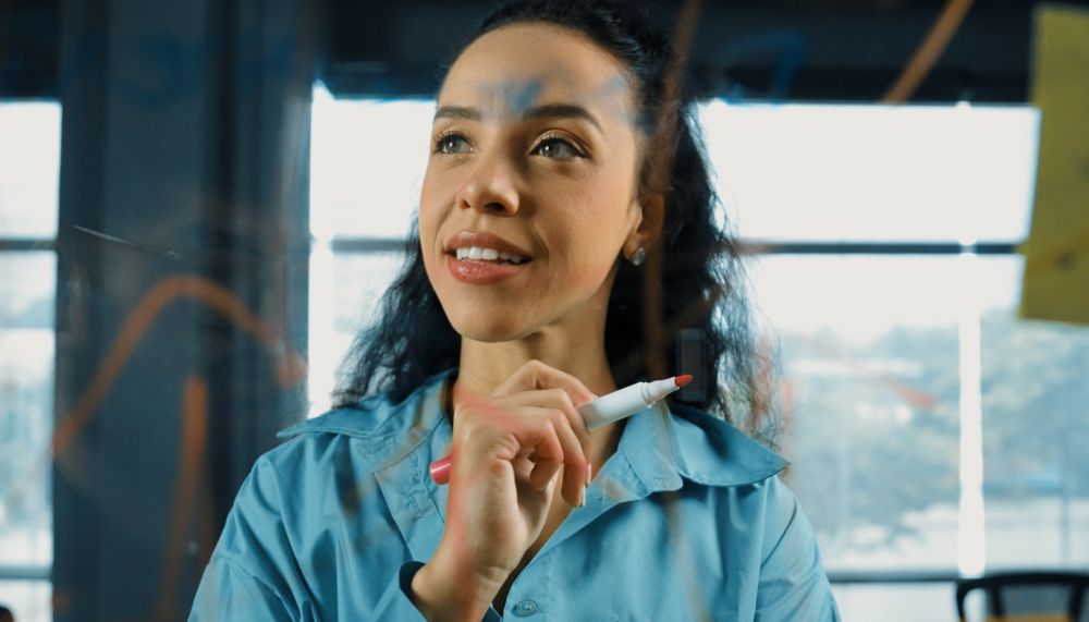 Durch ein beschriebenes  Glasboard ist eine junge Frau zu sehen, die einen Stift in der Hand hält.