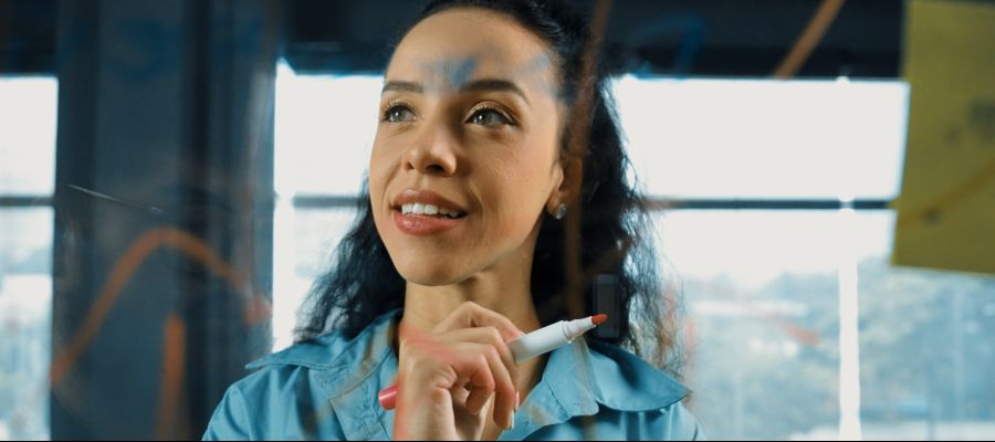 Durch ein beschriebenes Glasboard ist eine junge Frau zu sehen, die einen Stift in der Hand hält.