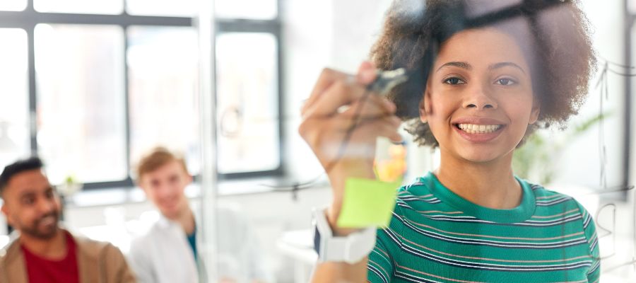 junge Frau, die auf Post-its an einer Glaswand schreibt. Im Hintergrund sitzen zwei weitere Personen