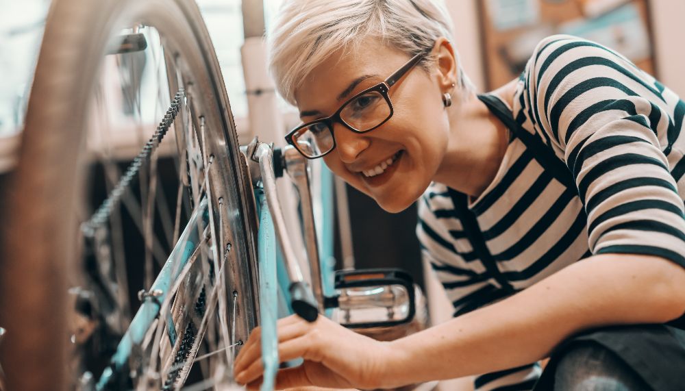 A woman is inspecting the rear tyre of a bicycle, which is propped up on its handlebars. 