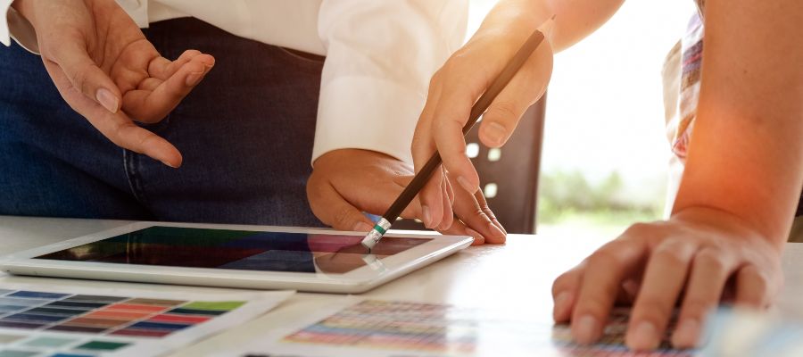 On voit les quatre mains de deux personnes qui gesticulent devant un plan et une tablette posés sur une table.