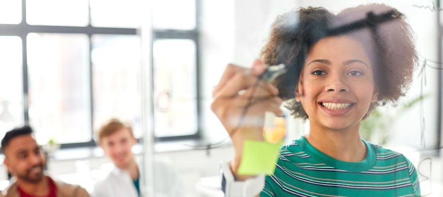 Une jeune femme qui écrit sur des Post-it collés sur une paroi vitrée. À l'arrière-plan, deux autres personnes sont assises.