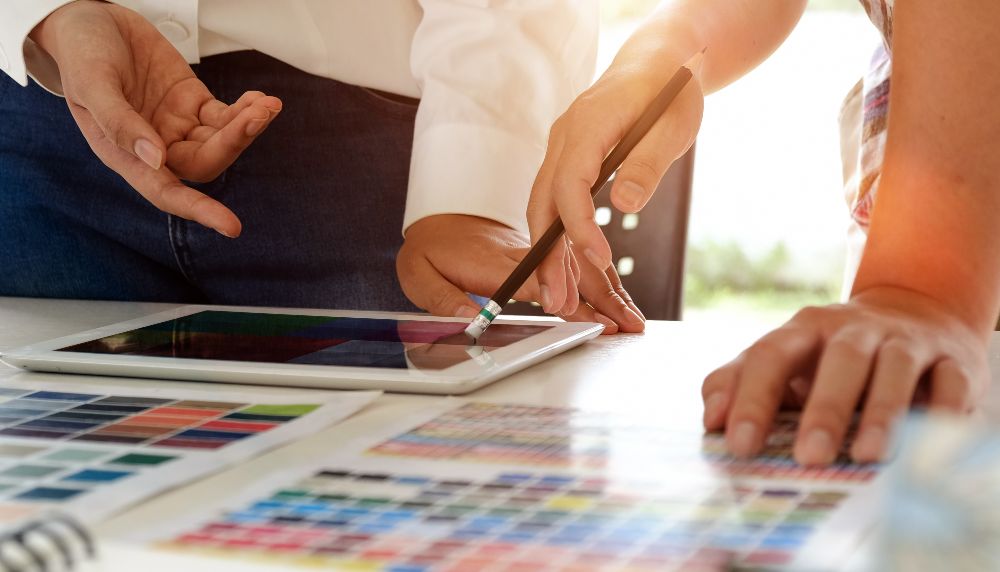  On voit les quatre mains de deux personnes qui gesticulent devant un plan et une tablette posés sur une table.