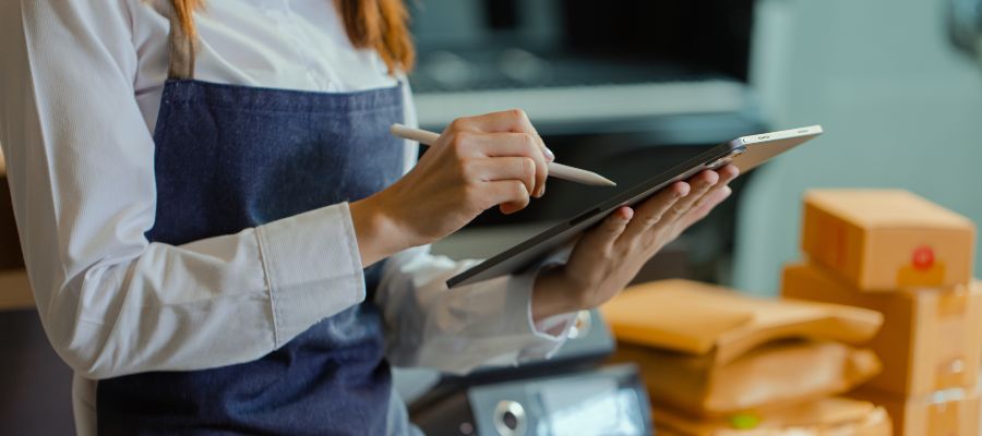 A woman wearing an apron and with red hair; only her hands are visible. Her left hand is holding a notepad, and her right hand is holding a pen, which she is using to tick something off on the notepad. In the background, there are files and small parcels.