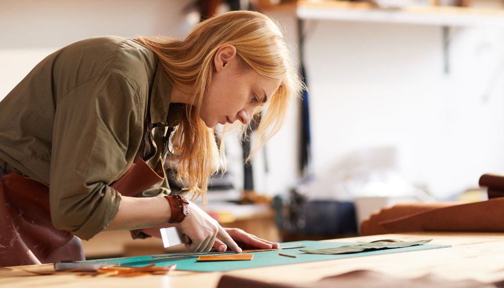 A young woman is cutting a piece of leather in a workshop.