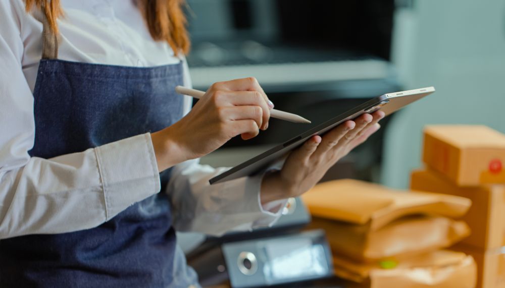A woman wearing an apron and with red hair; only her hands are visible. Her left hand is holding a notepad, and her right hand is holding a pen, which she is using to tick something off on the notepad. In the background, there are files and small parcels.