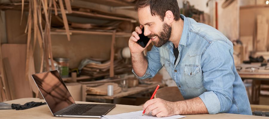 A man in a workshop is on the phone and jotting something down whilst looking at a laptop.