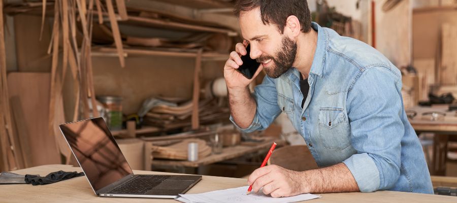 Un homme dans un atelier est au téléphone et prend des notes tout en regardant un ordinateur portable.