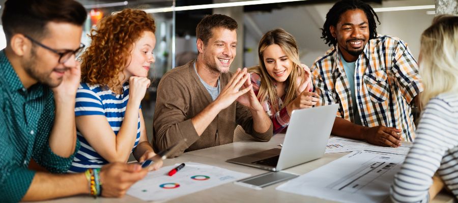 Des jeunes souriants sont debout autour d'une table sur laquelle se trouvent un ordinateur portable et des documents.