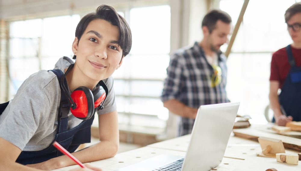 A young person wearing dungarees looks into the camera; in the background, two people can be seen out of focus at a workbench.
