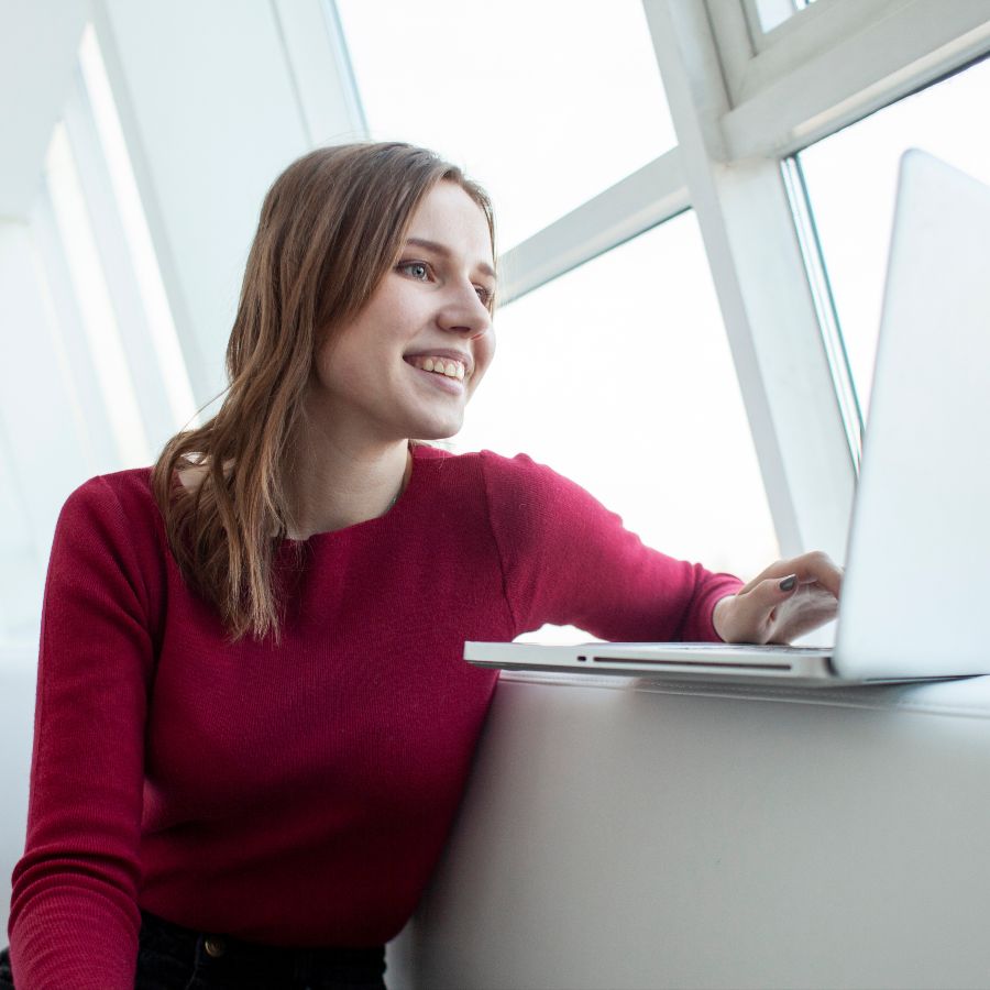 A woman in a red jumper is sitting at her laptop by a window.