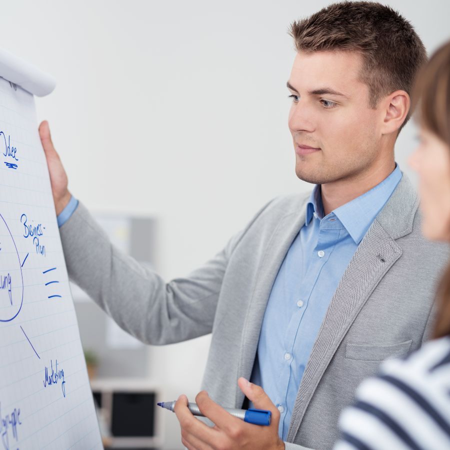 A young woman with dark hair is standing next to a flipchart, smiling at a man who can only be seen from behind in the foreground of the picture. 