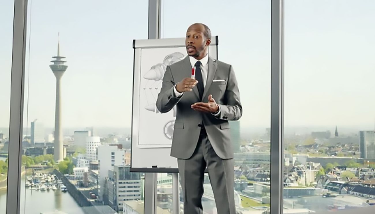 Ein Mann in einem grauen Anzug steht vor einem Flipchart und erklärt etwas. Im Hintergrund ist die Stadt Düsseldorf zu sehen.