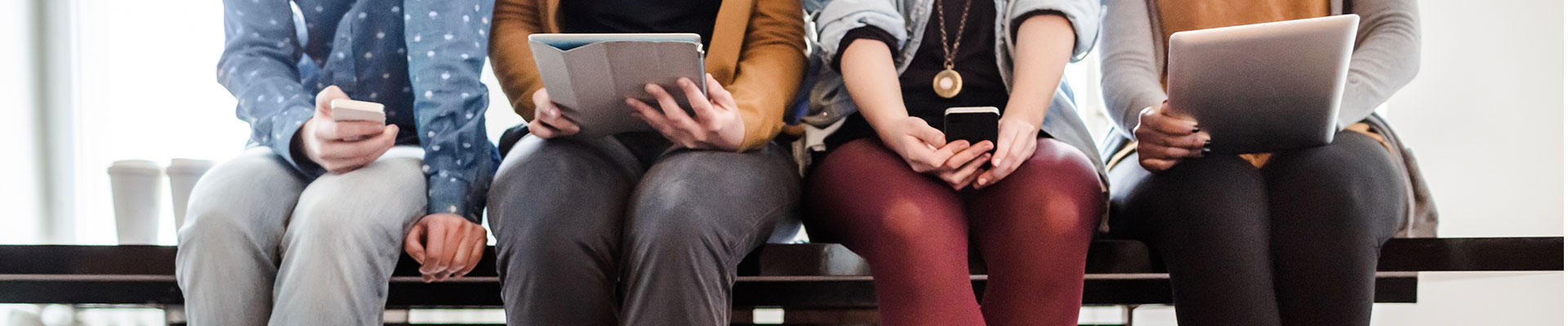 Image of knees of four people sitting side by side holding tablets or smartphones in their hands.