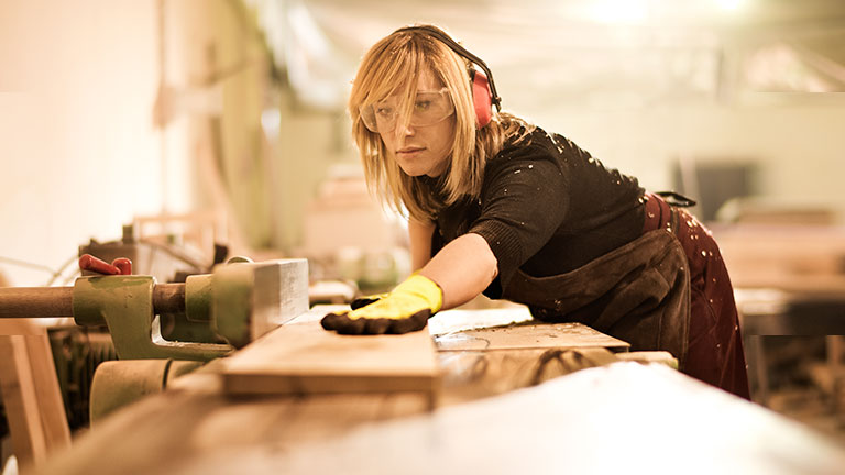 A woman wearing protective goggles and protective gloves works at a workbench.