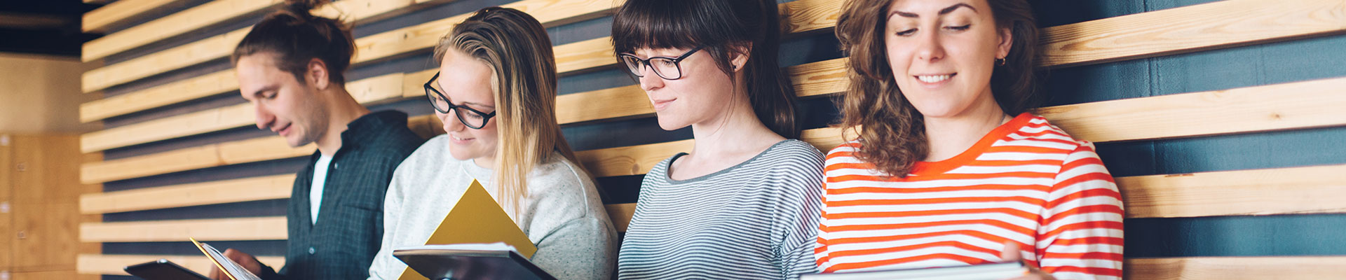 Young people sit leaning against a wooden wall and read in documents or tablets.