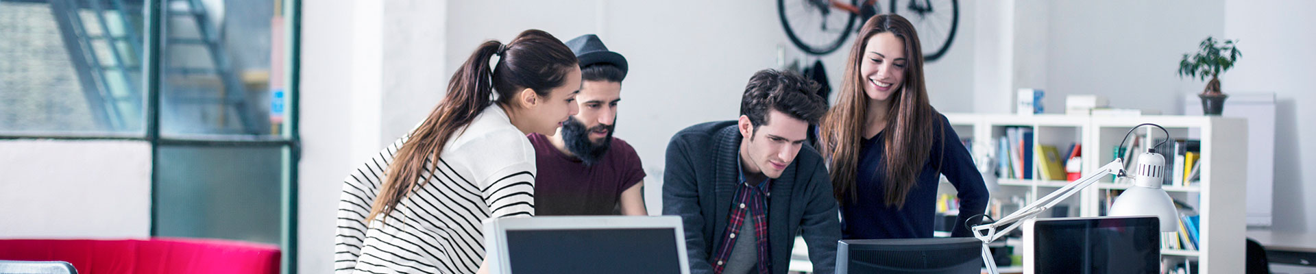 Four young people are staying in front of a desk and are looking at a screen. einen Bildschirm.