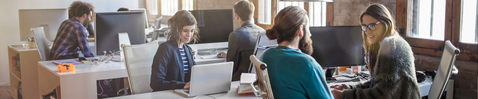 Young men and women work in an open-plan office with several desks.