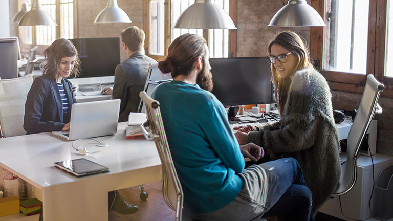 Young men and women work in an open-plan office with several desks.