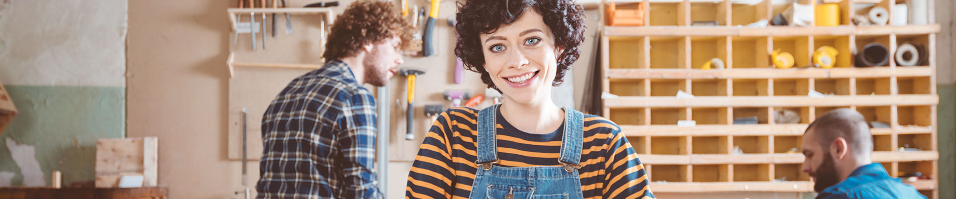 A young woman in dungarees stands in a workshop.