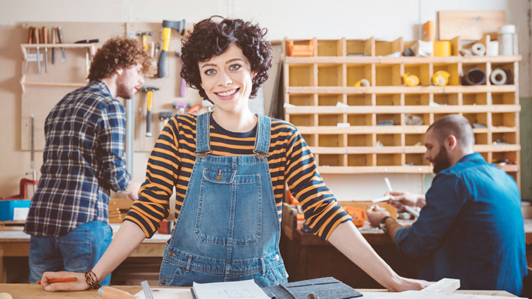 A young woman in dungarees stands in a workshop.