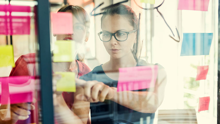 Two women look at post-its on a wall.