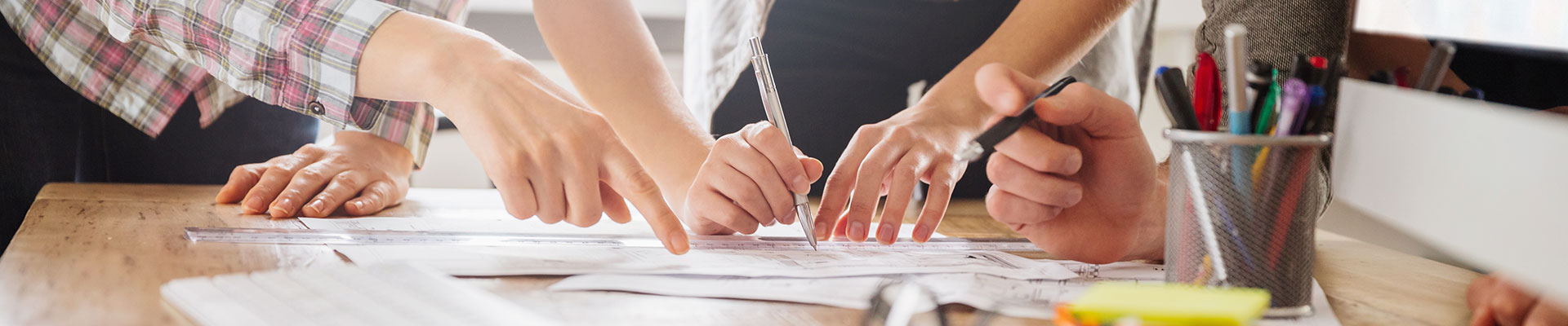 The hands of three persons point at or write on a paper on a desk.