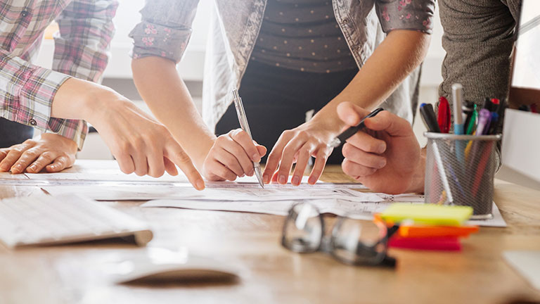 The hands of three persons point at or write on a paper on a desk.