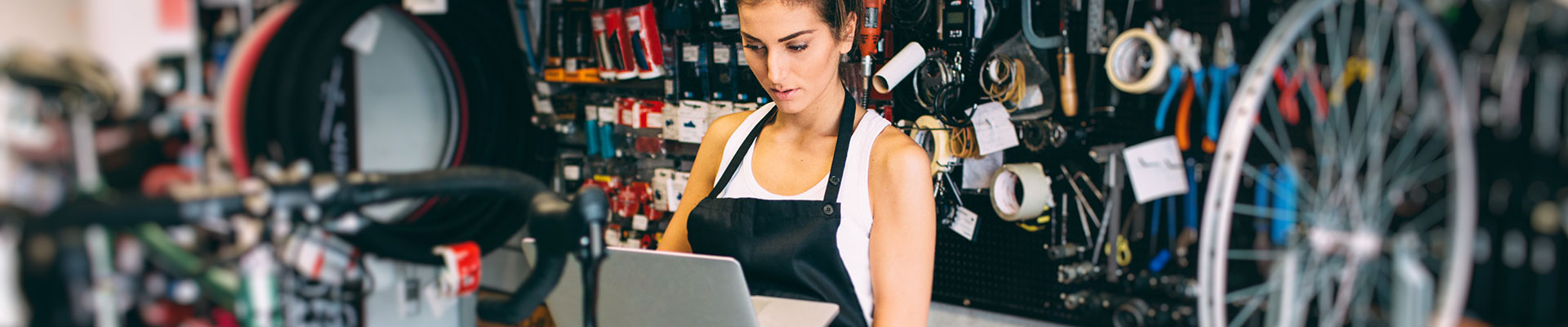 A young woman works in a bicycle repair shop at a laptop.