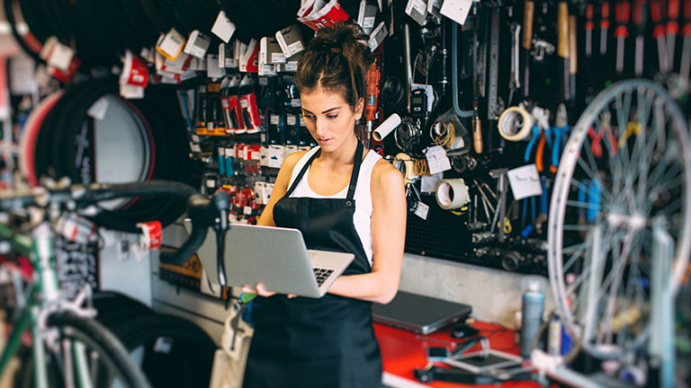 A young woman works in a bicycle repair shop at a laptop.