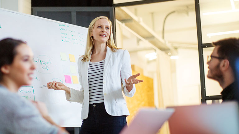 A woman stands in front of a whiteboard and gives a presentation. 