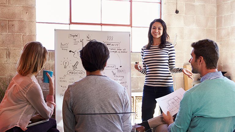 A young woman is presenting something to a group on a whiteboard. 