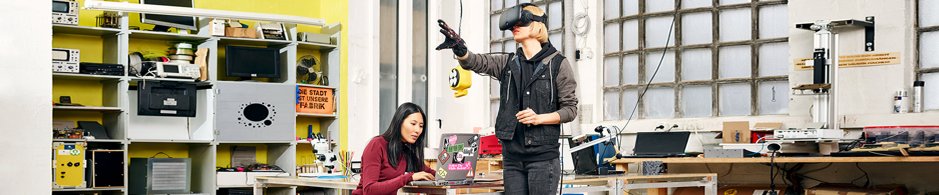 Zwei junge Frauen sitzen mit Laptop und VR-Brille in einer Werkstatt.