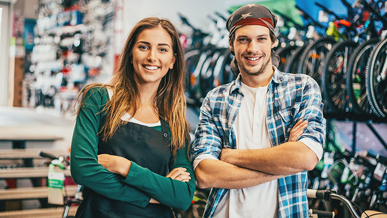 Jeune femme et jeune homme dans un magasin de bicyclettes, sourient à la caméra.