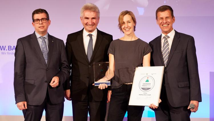 Three men and a woman stand in a row. The woman holds a certificate and an award.