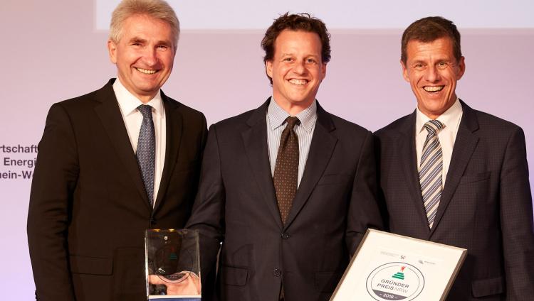 Three men stand in a row. The  middle holds a certificate. 