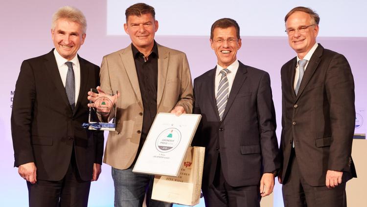 Four men stand in a row. The second from the left holds a certificate.
