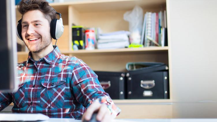 A young man with earphones sits in front of a computer. 