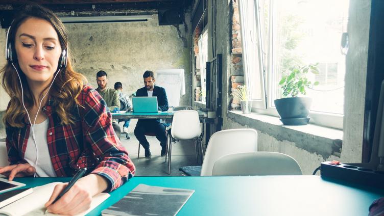 Une femme, avec des écouteurs, est assise à un bureau et écrit dans un carnet.