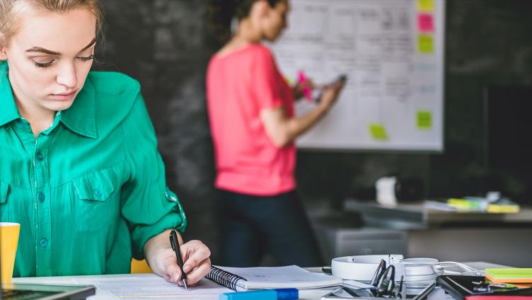 Une jeune femme est assise à un bureau, devant un ordinateur portable, et écrit des notes dans un carnet.