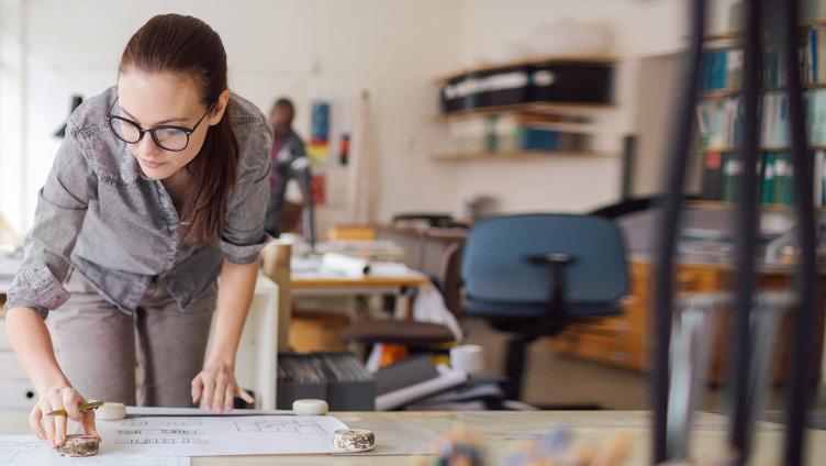 A young woman is leaning over a desk with drawings and sketches.