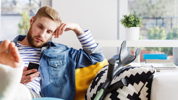 A young man with plaster leg lies on a sofa and look at his smartphone.