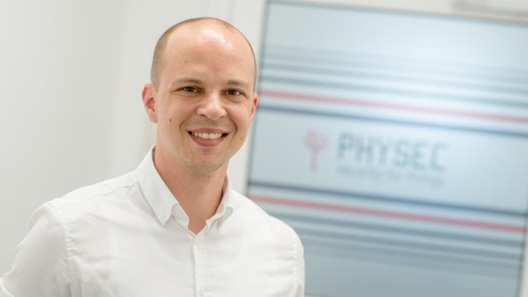 Dr. Heiko Koepke steht vor einer Wand mit dem Logo der Firma PHYSEC darauf